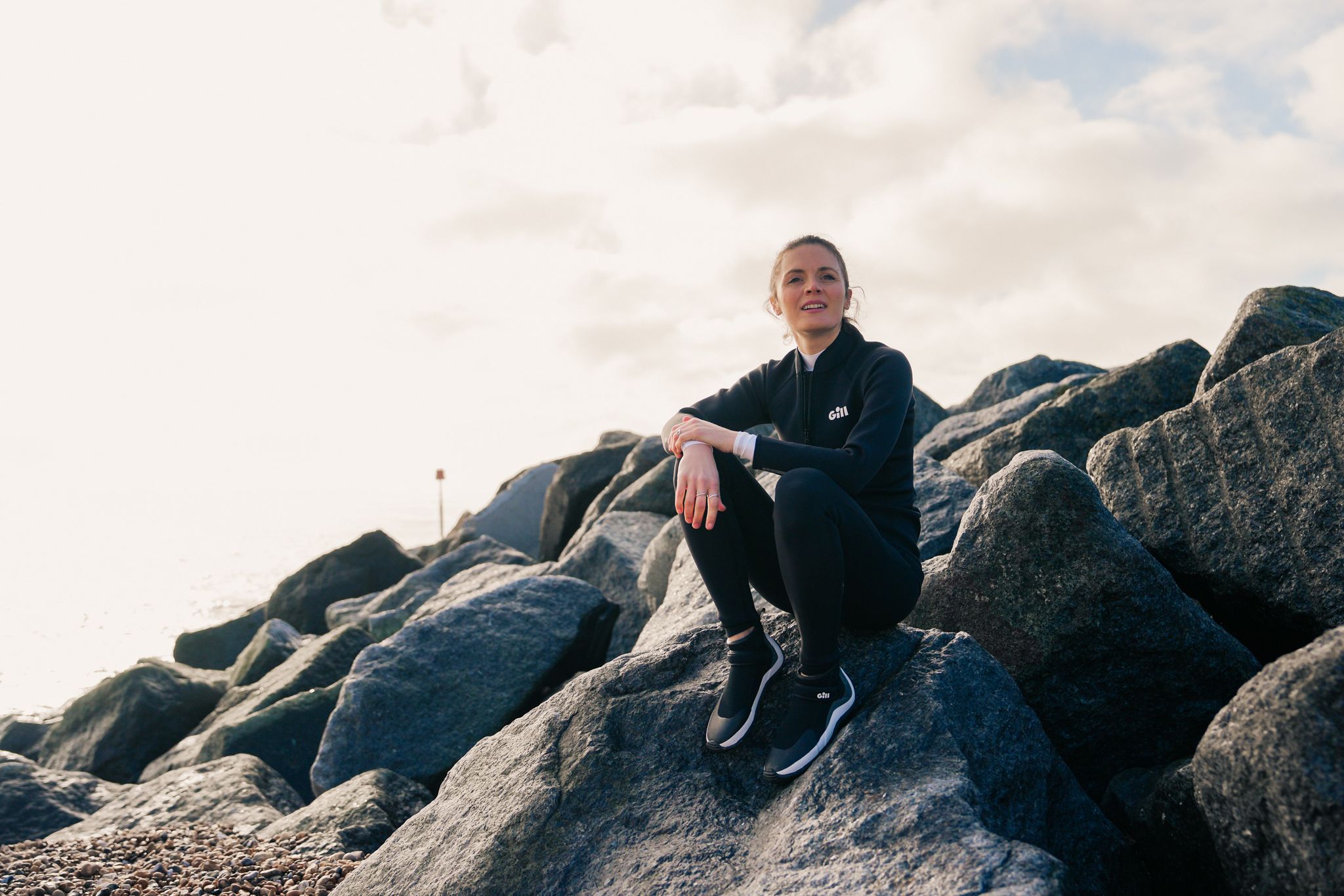 A woman sits a top some rocks in a wetsuit and looks out to the water.